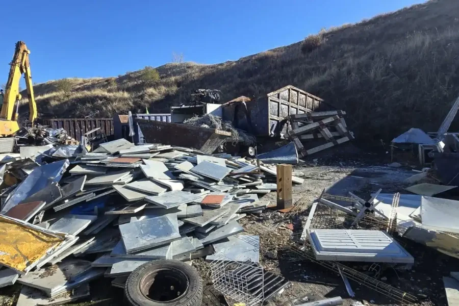 Un montón de escombros metálicos, electrodomésticos y materiales de desecho se encuentra en un depósito de chatarra al aire libre cerca de una colina de tierra junto a la Cañada. Una excavadora amarilla está aparcada a la izquierda, rodeada de neumáticos y objetos rotos, vestigios de la industria ilegal, bajo un cielo despejado.
