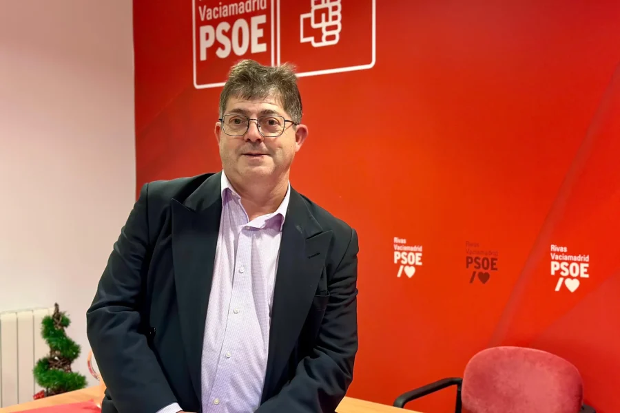 Alberto Cabeza, secretario general, con traje y gafas está de pie junto a una mesa en un despacho con paredes rojas que muestran los logotipos del PSOE de Rivas Vaciamadrid. Cerca, una silla roja y una pequeña planta completan la escena, en la que se pueden revisar documentos como un borrador automático.