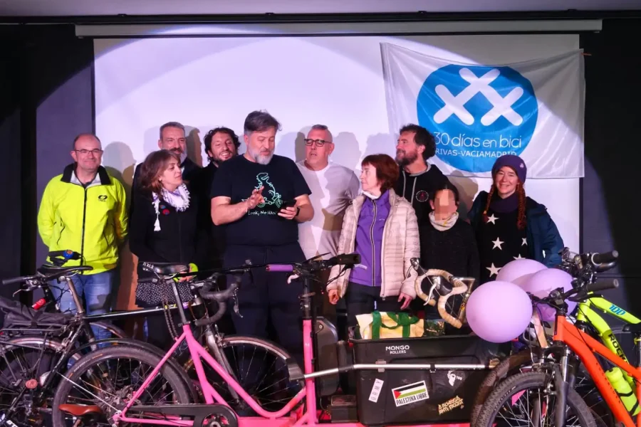Un grupo de ciclistas de pie en un escenario en Rivas detrás de varias bicicletas y material ciclista, con una pancarta de "30 días en bici" en la pared. Los individuos parecen estar escuchando al hombre de delante que lee en un papel.