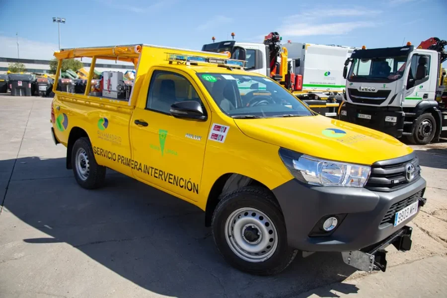 Una camioneta amarilla de Rivamadrid con la inscripción «Servicio de Primera Intervención» está aparcada en un aparcamiento junto a otros vehículos de servicio, probablemente preparándose para la campaña contra incendios de septiembre. La camioneta lleva las luces de emergencia encendidas, una baca y marcas de seguridad en los laterales.