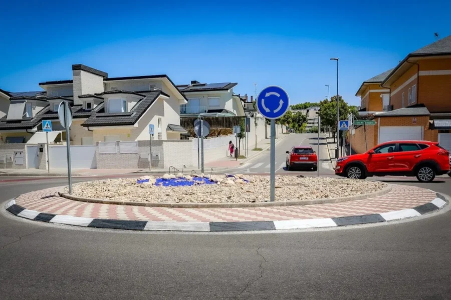 Una pequeña rotonda en la avenida Pilar Miró, con grava en el centro rodeada de ladrillos y baldosas, está rodeada de viviendas; se ven dos coches y un peatón, junto con señales de rotonda y de paso de peatones, lo que refleja un proyecto vecinal bien pensado.