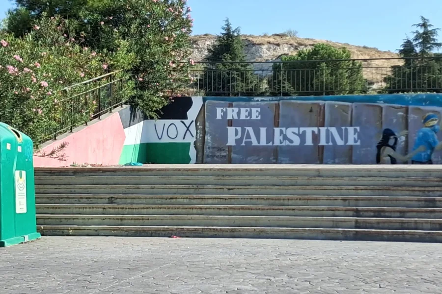 Una escalera de hormigón conduce a un mural en apoyo a Palestina con las palabras «FREE PALESTINE» y la palabra «VOX» sobre un fondo que recuerda a una bandera. A la izquierda de esta escena urbana, que recuerda a los espacios cercanos a Miguel Ríos, se aprecian árboles, un contenedor de reciclaje y referencias a «Convocatoria en Rivas».