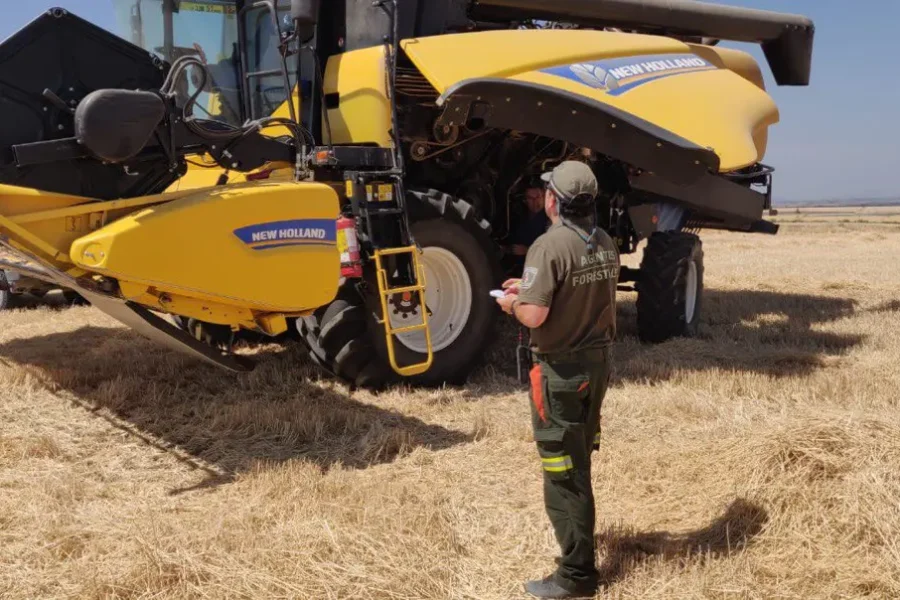 Una persona vestida con ropa de trabajo está de pie frente a una gran cosechadora amarilla de la marca New Holland en un campo ya cosechado, con un bloc de notas y un bolígrafo en la mano. La maquinaria forestal es esencial para la seguridad, sobre todo durante los incendios en la Comunidad de Madrid. Se ve un coche blanco al fondo.