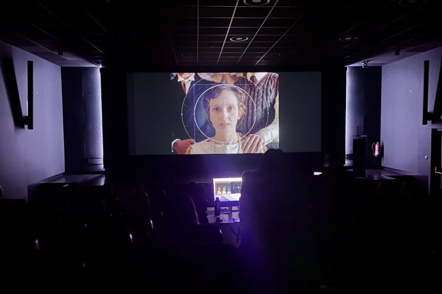 Una sala de cine poco iluminada de la Escuela de Cinematografía de la Comunidad de Madrid proyecta una película en la que aparece una mujer vestida de época. El público está sentado en la sombra, con la luz de un panel de control que ilumina parte de la sala.