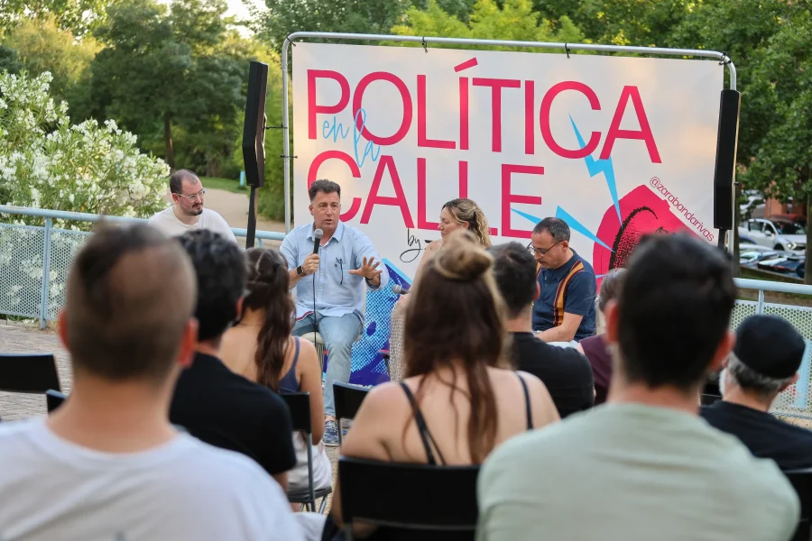 Cuatro personas forman parte de una mesa redonda frente a un público al aire libre, hablando bajo un cartel que reza «POLÍTICA en la CALLE». Entre ellas se encuentran Vanessa Millán y Pedro del Cura, que debaten sobre el modelo de ciudad de Rivas con árboles y una valla al fondo.