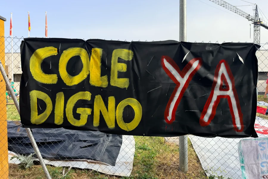 Una pancarta negra fijada a una valla del CEIP Mercedes Vera muestra las palabras COLE DIGNO en amarillo y YA en grandes letras rojas y blancas, con materiales de construcción, una grúa y obras en curso visibles en el fondo.