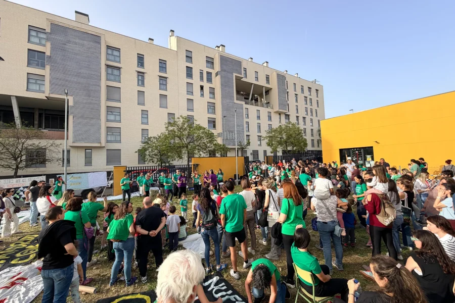 Un nutrido grupo de personas, muchas de ellas con camisetas verdes, se reúnen al aire libre en una zona de césped frente a un moderno edificio de apartamentos y un muro amarillo durante un encierro diurno para protestar por el retraso de las obras en el CEIP Mercedes Vera.