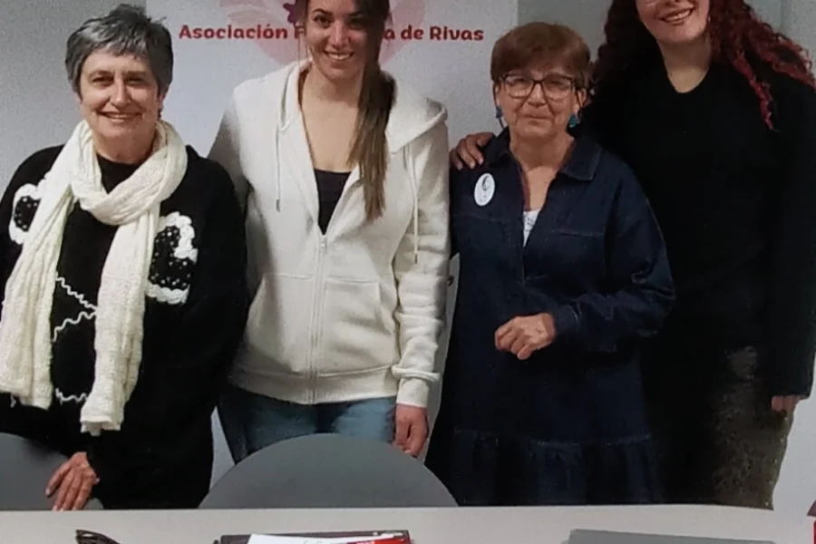Cuatro mujeres de la nueva Junta Directiva de la Asociación Feminista 13Rosas están juntas delante de una pancarta de 13Rosas. Sonriendo a la cámara, están reunidas detrás de una mesa con un portapapeles, llaves, un cordón y un ordenador portátil.