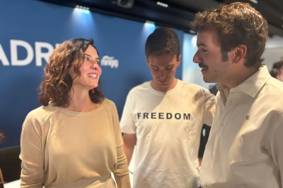 Tres personas conversan en un interior. La mujer de la izquierda sonríe con un top beige. El hombre del centro, con una camiseta blanca de LIBERTAD, es Jesús Martínez Caballero, de NNGG Madrid. El hombre de la derecha lleva una camiseta clara.