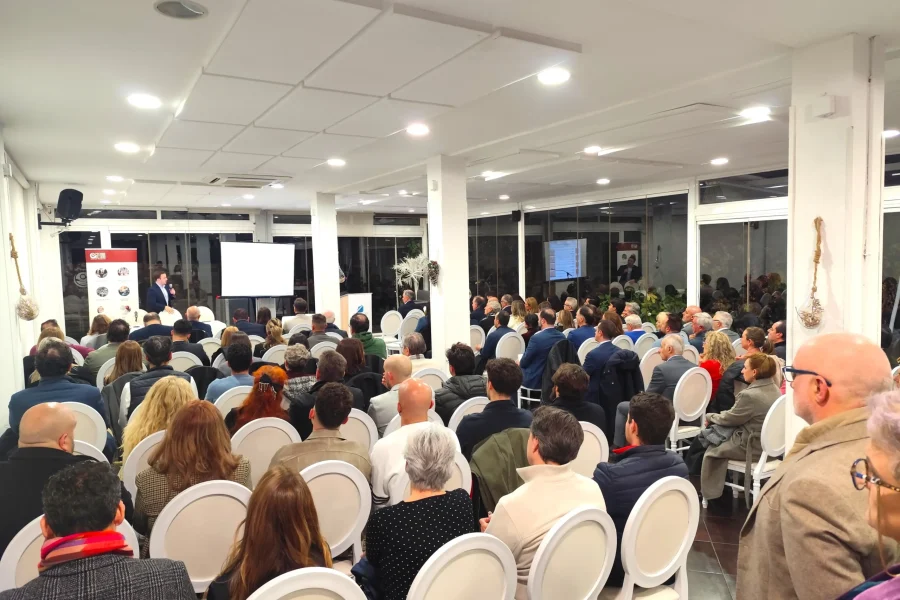 Un nutrido grupo de empresarios se sienta en sillas blancas en una sala de conferencias muy iluminada, frente a un escenario donde habla un presentador de ASEARCO cerca de una pantalla de proyección. La sala tiene paredes blancas, pilares y grandes ventanales durante el encuentro navideño.