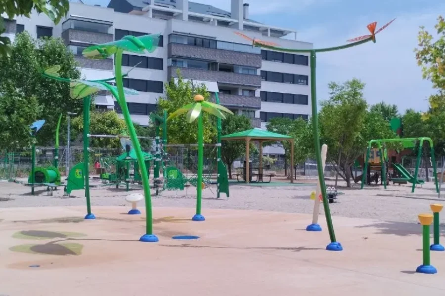 Parque infantil al aire libre con estructuras de juego de colores verde y azul, con juegos de agua, situado en una zona de arena y rodeado de árboles, con edificios residenciales al fondo. En el Parque del 8M también se pueden ver columpios y estructuras para trepar.