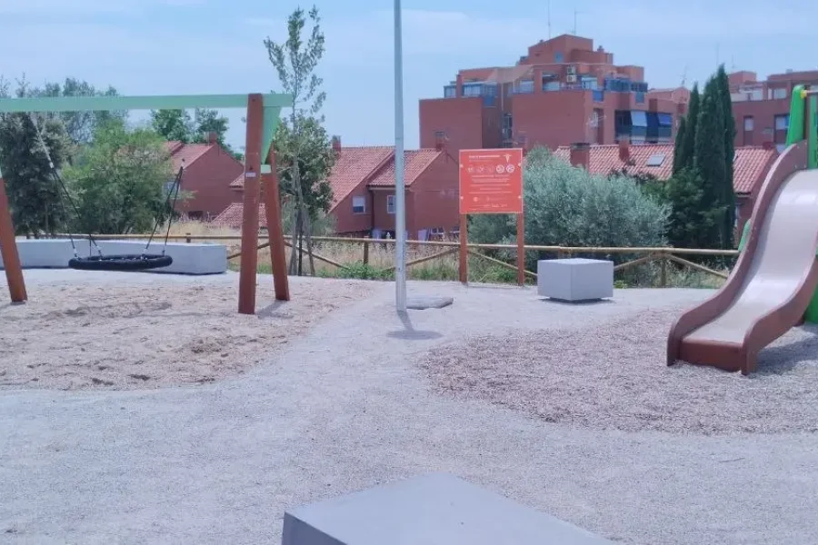 Un parque infantil con un columpio y un tobogán sobre suelo de grava ofrece espacios para los niños en Rivas. Al fondo se ve un cartel naranja, mientras que detrás de una valla de madera se divisan edificios residenciales y árboles. El cielo está parcialmente nublado.