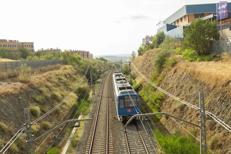 Un tren de cercanías azul circula por una vía doble a través de un desmonte seco y cubierto de hierba entre colinas, pasando junto a viviendas y edificios a ambos lados bajo un cielo despejado, con cables eléctricos que se extienden por encima.