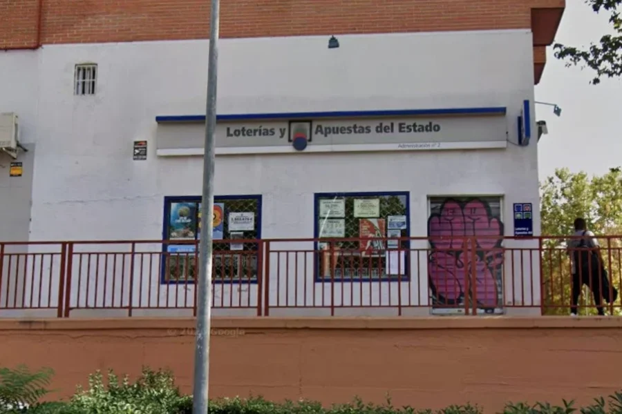 Una oficina de Loterías y Apuestas del Estado en Rivas, con una barandilla metálica roja en la fachada y una persona que pasa junto al edificio.