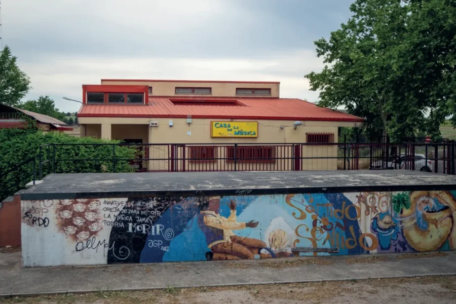 Un edificio de una sola planta con tejado rojo y un cartel amarillo en el que se lee Casa de la Música; enfrente, una pared pintada con grafitis que evoca uno de los cuatro sitios donde hacer (y escuchar) música, además de una zona vallada con árboles a la derecha.