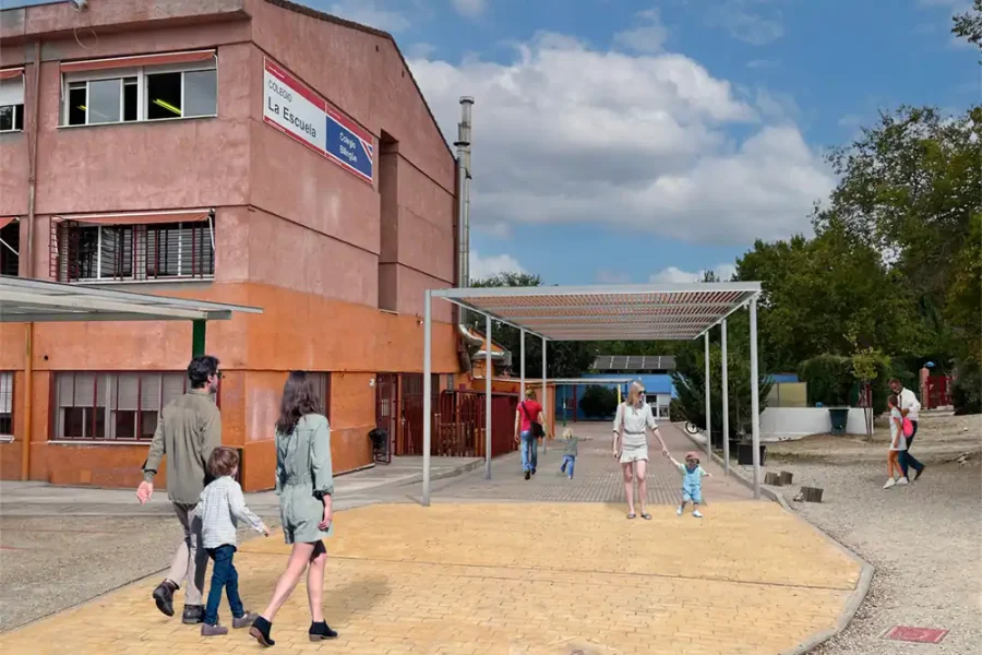 Varios adultos y niños pasean por el exterior de un colegio público de Rivas, en el que destacan sus paredes rojas y marrones. Algunos niños juegan bajo las pérgolas, mientras el cielo está parcialmente nublado sobre las pasarelas adoquinadas y el descampado.