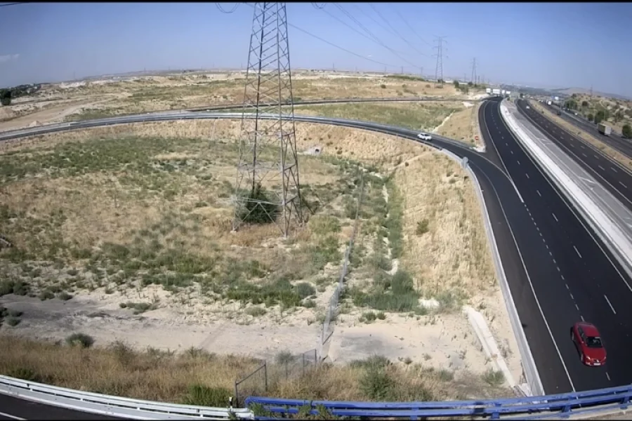 Vista aérea del enlace de la autopista M-50 cerca de Rivas, con una rampa de acceso en bucle que rodea hierba seca y una alta torre eléctrica; varios vehículos circulan por la carretera bajo un cielo despejado.
