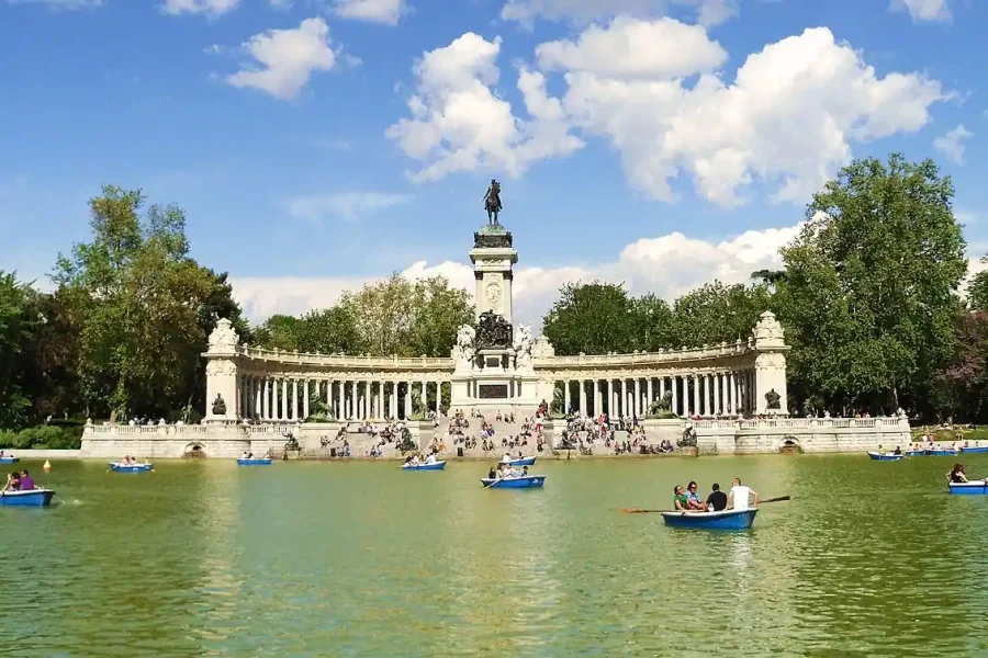 La gente rema en barcas azules en un gran lago de El Retiro, frente a un gran monumento de piedra con columnas y una estatua en el centro, rodeado de árboles y un cielo parcialmente nublado, que ofrece accesibilidad a todos los visitantes.