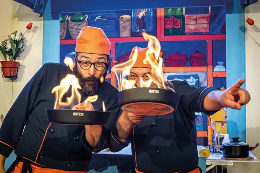 Dos cocineros, con gorros naranjas y uniformes negros, sostienen sartenes de las que brotan llamas en Festaclown 2026; uno de ellos señala hacia delante, mientras que al fondo se ven unas estanterías de cocina de colores.