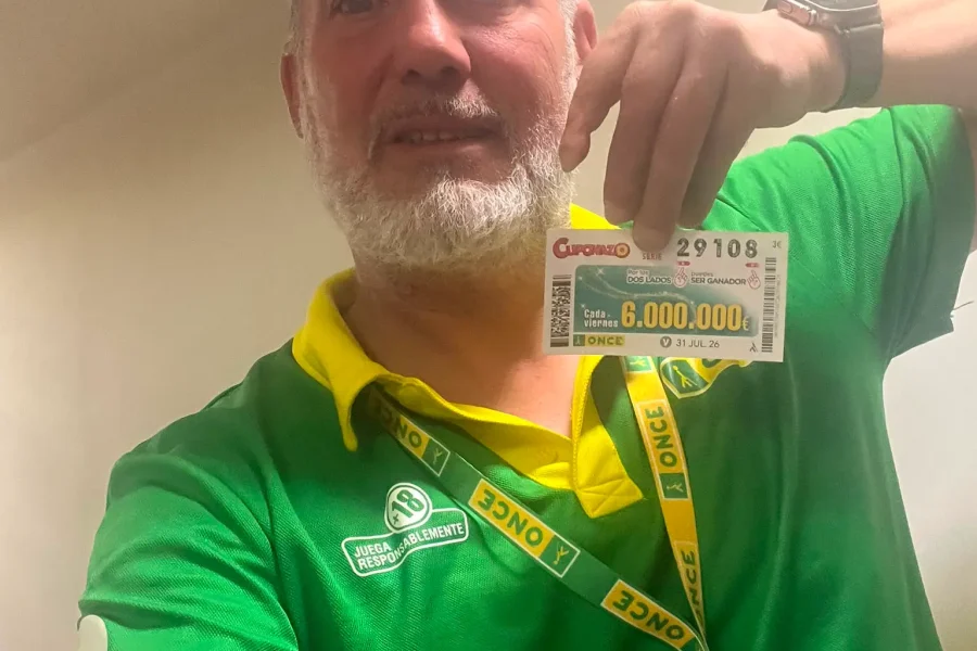 Un hombre con una camiseta verde y amarilla de la ONCE sostiene un boleto de lotería. Lleva barba, lleva una insignia y se encuentra en el interior, posiblemente preparándose para el «Cuponazo ONCE» o el sorteo de los «premios Rivas».