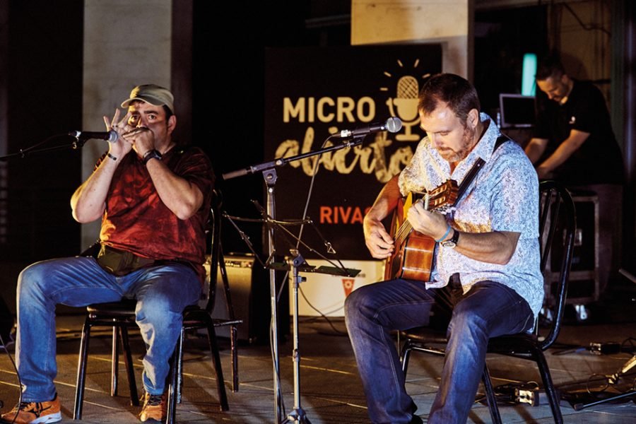 Dos músicos tocan la armónica y la guitarra en el escenario del Micro Abierto de Rivas, con el cartel del programa al fondo