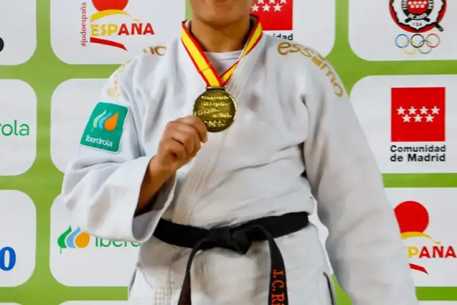 Nisrin Bousbaa, atleta de judo con gi blanco y cinturón negro, sostiene su medalla de oro y sonríe ante un telón de fondo con logotipos y texto en español tras ganar su tercer oro nacional.