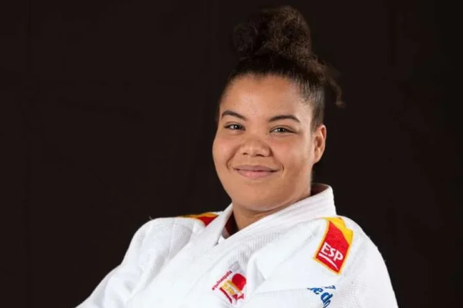 Nisrin Bousbaa, vestida con un uniforme blanco de judo y un cinturón negro, sonríe con los brazos cruzados delante del logotipo de la Federación Internacional de Judo.