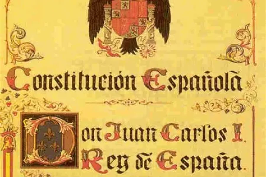 Una página decorativa con texto ornamentado y un escudo real en la parte superior, titulada "Constitución Española", en referencia al rey Juan Carlos I de España, destaca el marco constitucional y presenta una caligrafía y unos bordes elaborados.