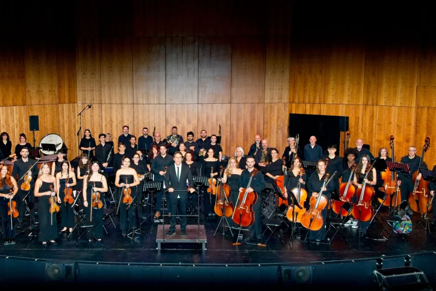 Una orquesta completa, compuesta por músicos de cuerda, viento-metal, viento-madera y percusión, posa en el escenario vestida de concierto con sus instrumentos, lista para interpretar música de cuerda o selecciones de ópera y zarzuela. El escenario presenta paneles de madera al fondo.