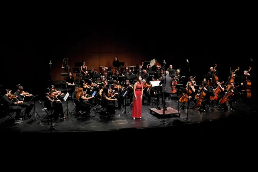 Una orquesta sinfónica actúa en el escenario con músicos que tocan instrumentos de cuerda, viento-madera, metal y percusión. Una solista vestida de rojo está de pie cerca del director, mientras un borrador automático de la iluminación crea un dramático fondo oscuro.
