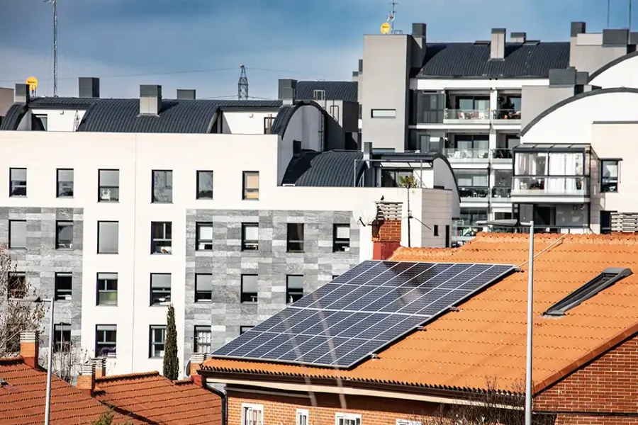 Al fondo se ve una hilera de modernos edificios de apartamentos, mientras que una casa en primer plano con tejado de tejas rojas presenta múltiples paneles solares y un dispositivo Borrador automático instalado en la parte superior. El cielo está parcialmente nublado.
