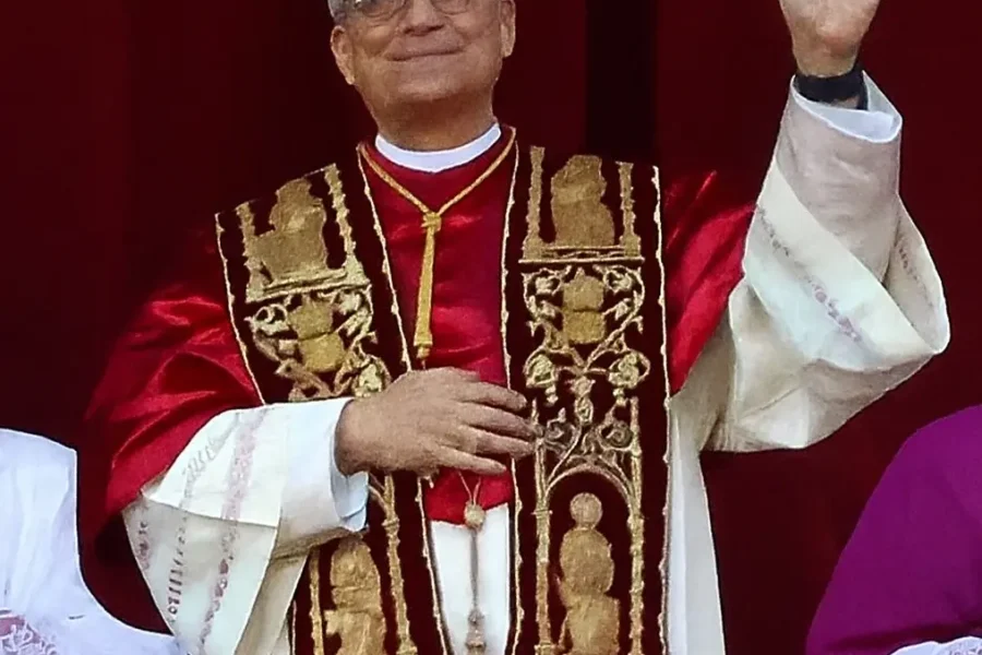Un clérigo de la Iglesia Católica, ataviado con ornamentadas vestiduras religiosas, permanece de pie delante de una cortina roja, sonriendo y levantando la mano derecha en un gesto, con otras personas parcialmente visibles a ambos lados.