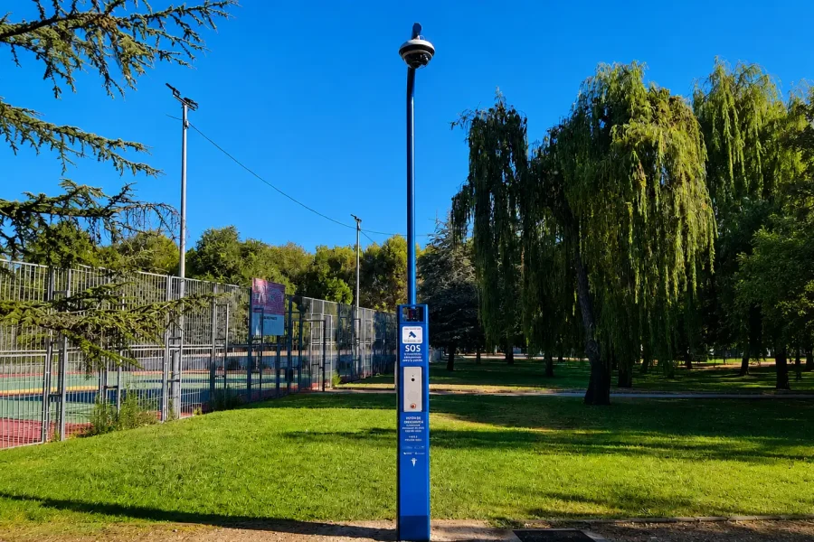 Una cabina azul de llamadas de emergencia, uno de los «SOS postes» de Rivas, equipada con una cámara de seguridad, se encuentra en una zona con césped cerca de una pista de tenis y de unos árboles, en un parque bajo un cielo azul y despejado, lo que aporta mayor seguridad y apoyo frente a la violencia machista.