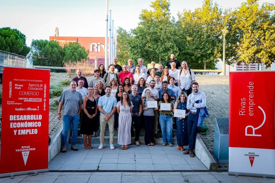 Un grupo de unas 35 personas posa al aire libre en unas escaleras; algunas sostienen certificados que reconocen sus proyectos o empresas. A ambos lados, dos pancartas rojas con los lemas «Rivas Emprende» y «Desarrollo Económico y Empleo» resaltan el espíritu de los Premios de Rivas. Al fondo se ven árboles y un edificio de ladrillo.