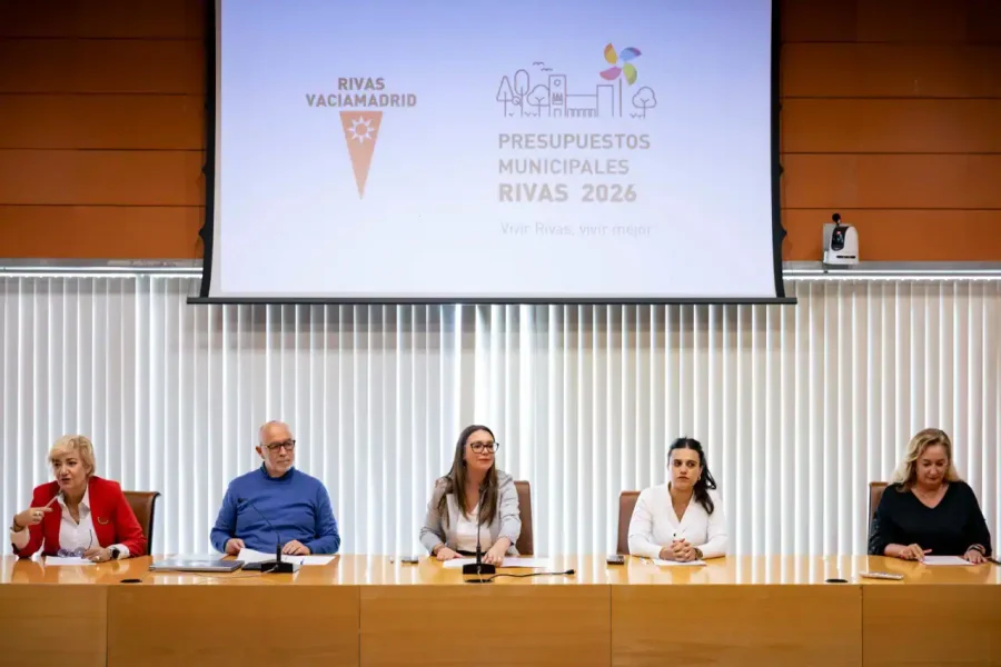 Cinco personas se sientan a una larga mesa de madera en una sala de conferencias. Detrás de ellos, una gran pantalla muestra "Presupuestos Municipales Rivas 2026", destacando iniciativas clave como la climatización escolar, con logotipos y elementos gráficos de Rivas Vaciamadrid.
