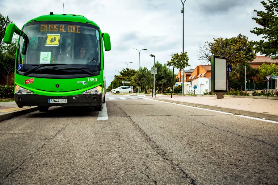Un autobús escolar verde con la inscripción «EN BUS AL COLE», que forma parte de las rutas escolares 2026/2027, está aparcado en una parada de autobús en una calle de las afueras. Las casas, los árboles, la acera y un cielo nublado conforman el fondo.