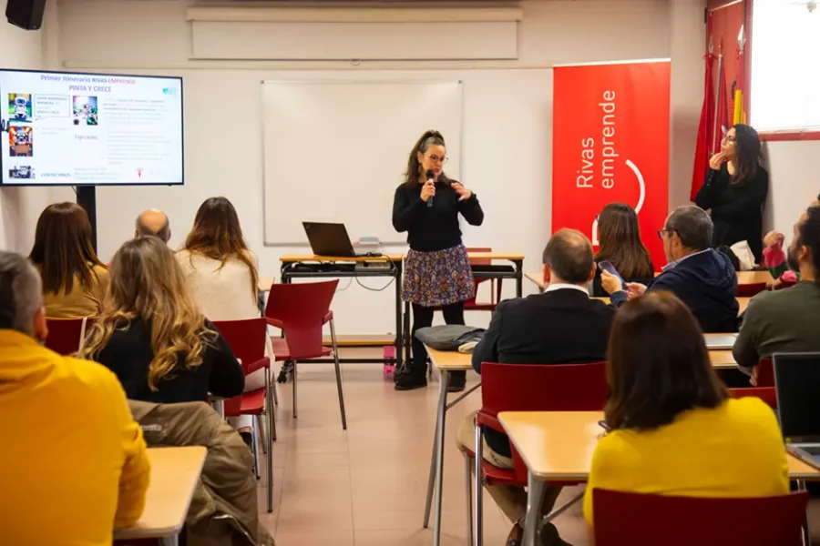 Una mujer está de pie al frente del aula hablando por un micrófono, con una presentación —titulada «Borrador automático»— proyectada en la pantalla que hay detrás de ella. Los participantes, sentados, miran hacia delante y escuchan. A la derecha, una pancarta roja reza «Rivas emprende».