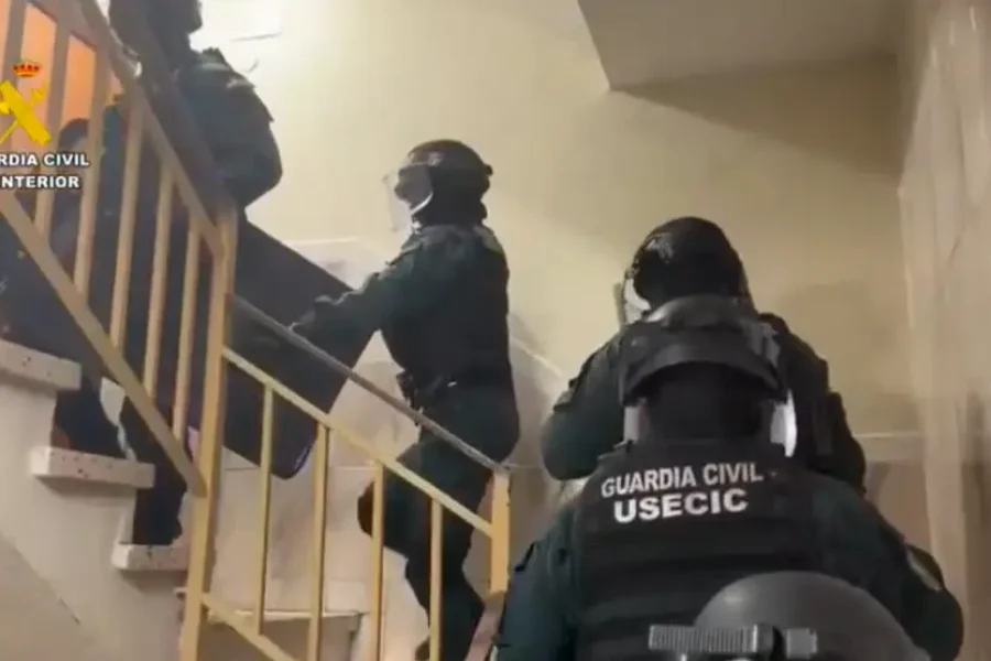 Cuatro agentes de policía, equipados con material táctico y cascos, suben por una escalera. En sus uniformes se lee la inscripción «GUARDIA CIVIL USECIC», lo que indica su participación en operaciones especializadas contra los ladrones de viviendas. La escena se desarrolla en el interior de un edificio en Rivas, con paredes de color beige y un pasamanos amarillo.