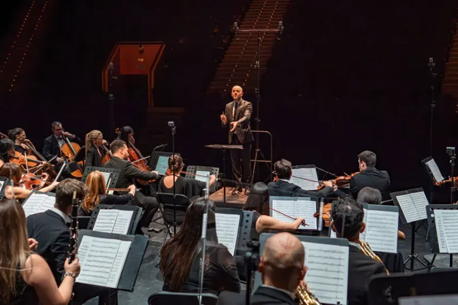 Un director de orquesta, de pie sobre un podio, dirige una orquesta de músicos que tocan varios instrumentos en el escenario, con partituras en atriles y filas de asientos vacíos visibles en el fondo poco iluminado.