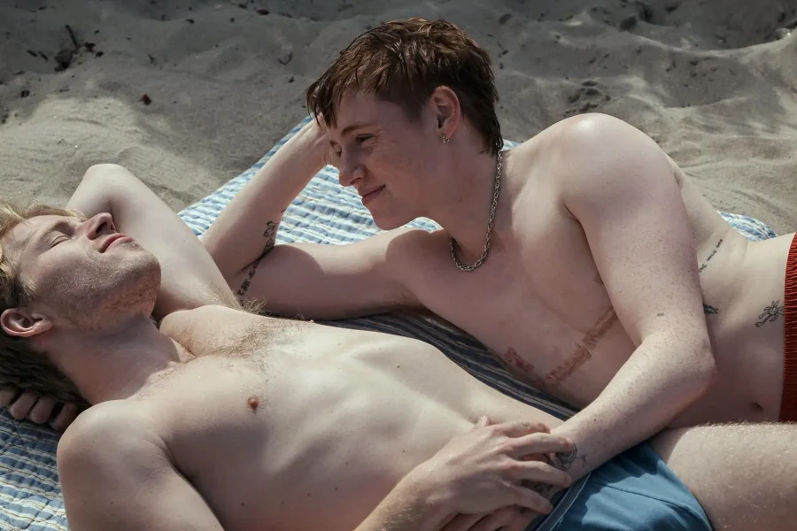 Dos descamisados tumbados en una toalla de rayas en la playa, frente a frente. Su momento de relajación evoca escenas de QueerCineMad, el Festival Internacional de Cine de la Comunidad de Madrid, con la arena rodeando su conexión iluminada por el sol.