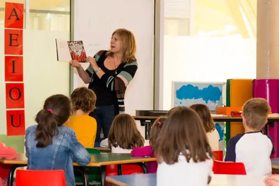 Un profesor, de pie delante de una clase, sostiene un libro y lee a los alumnos sentados en las mesas. A la izquierda, en una pizarra, hay grandes tarjetas rojas con las vocales, creando un ambiente similar al de los talleres creativos.