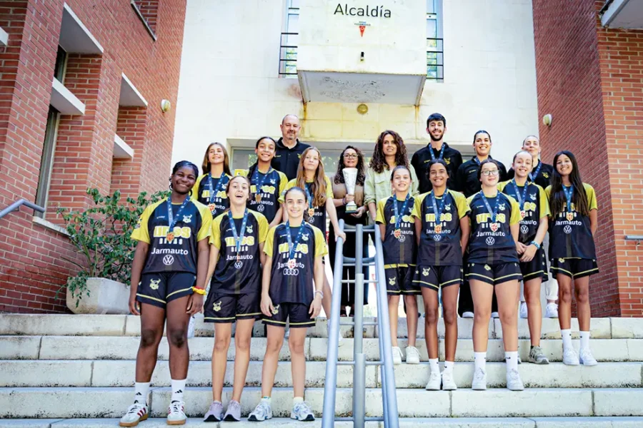 Un equipo de baloncesto femenino posa en unas escaleras al aire libre frente a un edificio, con varios adultos detrás de ellas. Las jugadoras, que representan al Uros de Rivas, llevan uniformes negros y amarillos y medallas. Sobre el grupo se lee un cartel que dice «Alcaldía», en el que se celebra su reciente éxito en la cantera en su camino hacia un título nacional.