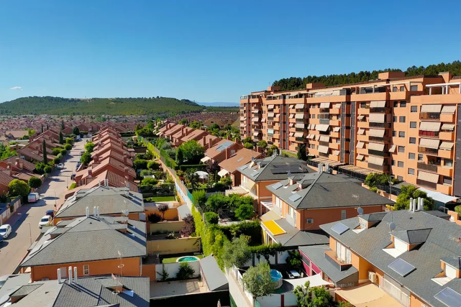 Vista aérea de un barrio residencial con hileras de casas, céspedes verdes y un edificio de apartamentos alto a la derecha. Al fondo se divisan colinas y un cielo azul claro. La zona destaca por su cuidada planificación urbanística, con bonificaciones especiales en el IBI disponibles para las familias numerosas de la comunidad.