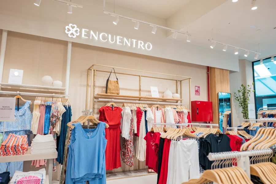 Interior de una tienda de ropa con percheros llenos de prendas de mujer de colores vivos, entre las que se incluyen vestidos y tops, cuidadosamente expuestas. En un estante alto hay un bolso marrón. El letrero de la tienda, con las palabras «Encuentro Modas» en letras blancas, se encuentra en el Centro Comercial H2O de Madrid.
