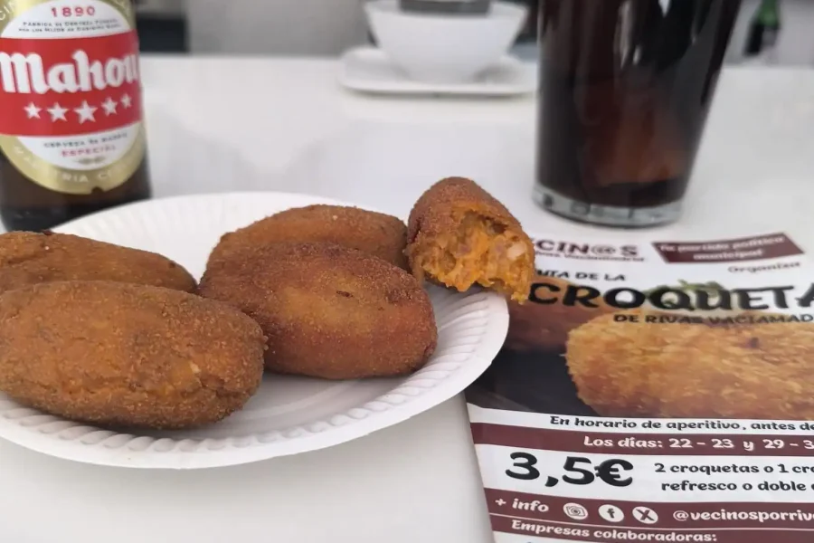 Un plato blanco con cuatro croquetas, una de ellas parcialmente comida, está sobre un mostrador junto a una botella de cerveza Mahou y un vaso de gaseosa oscura. Al fondo se ve un folleto de Borrador automático y otros objetos.