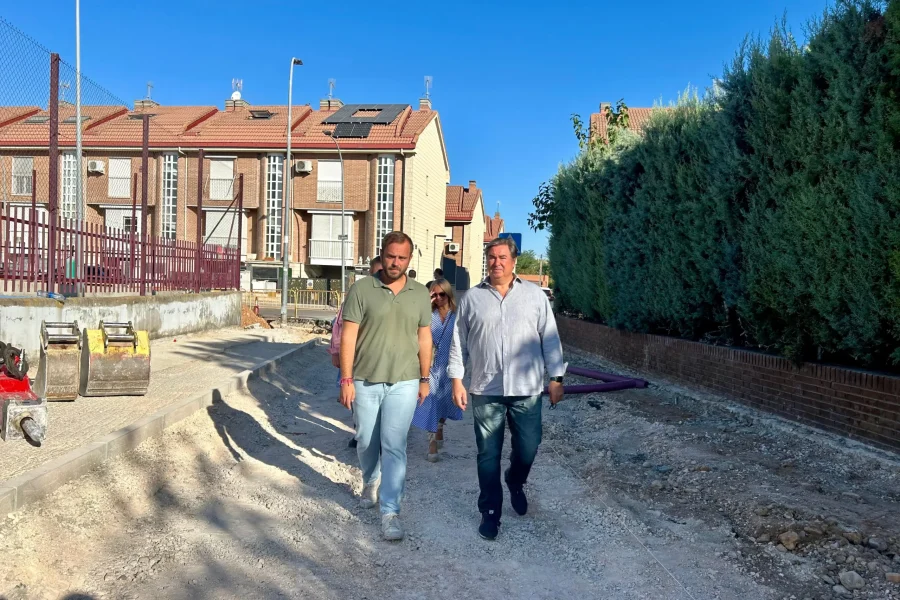 Dos hombres caminan codo con codo por un sendero de tierra en una obra de renovación de calles en Arganda del Rey, Comunidad de Madrid, pasando junto a casas y un seto alto con maquinaria de construcción cerca bajo un cielo azul despejado.