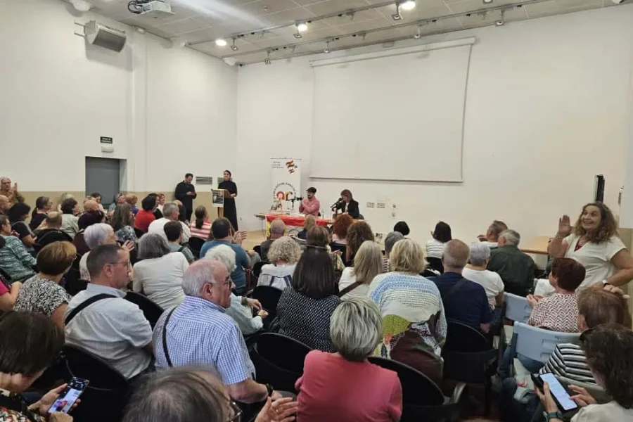 Una multitud se sienta frente a un panel de ponentes en una conferencia en una sala bien iluminada. Silvia Intxaurrondo presenta una novela desde el podio, mientras otra persona a la derecha gesticula con una mano levantada. En la pared del fondo hay una pantalla de proyección.