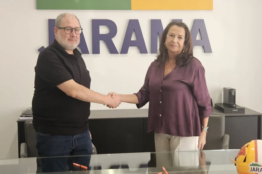 Un hombre y una mujer se dan la mano en un despacho frente a un cartel en la pared en el que se lee JARAMA, destacando su conexión con el deporte local en Rivas. Una mesa de cristal con marcadores rojos y un casco están en primer plano mientras ambos miran hacia la cámara.
