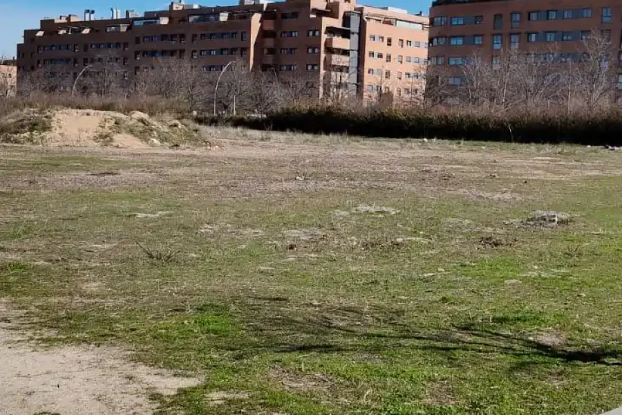 Una hilera de edificios de apartamentos marrones se alza tras un descampado cubierto de hierba y parcialmente sucio, con una acera asfaltada y una valla en primer plano y un cielo azul despejado en lo alto.