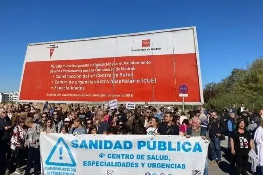 Un grupo numeroso de personas frente a una valla sostiene una pancarta en la que se lee "Sanidad Pública, 4º Centro de Salud, Especialidades y Urgencias." La valla publicitaria de detrás hace referencia a la construcción de un cuarto centro de salud.